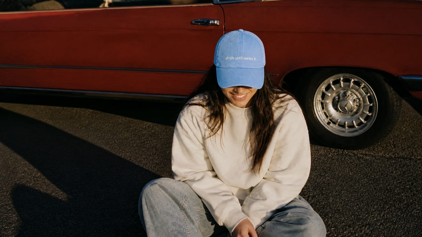 woman in a light blue single until series b cap sitting against a red convertible